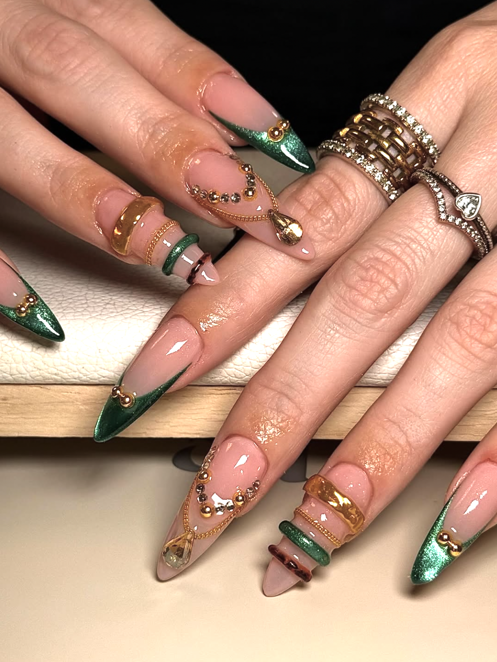 Long pointed almond nails with a nude base, deep emerald green tips, and various 3D gold bangle textures and chains.