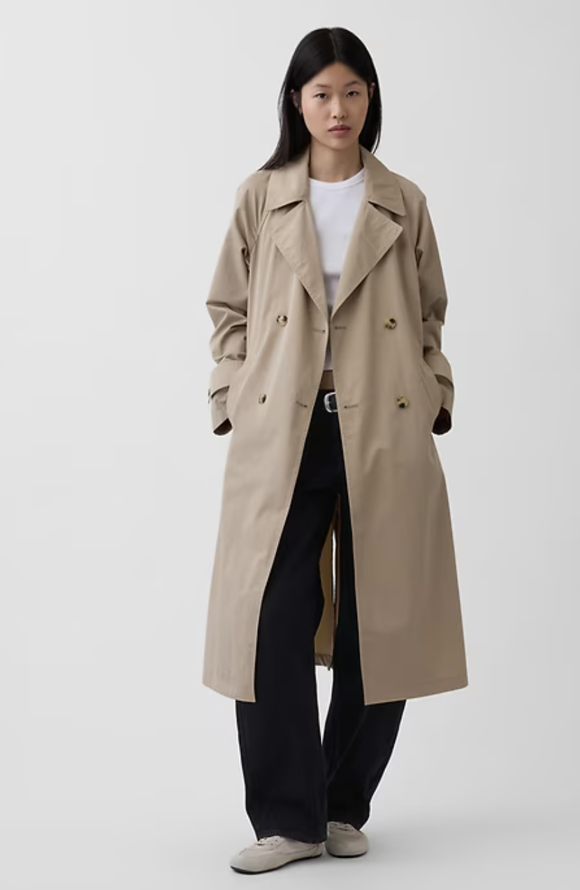 A person wearing a classic tan, long-line trench coat styled over a white t-shirt and dark trousers, featuring a belted waist and traditional lapels.