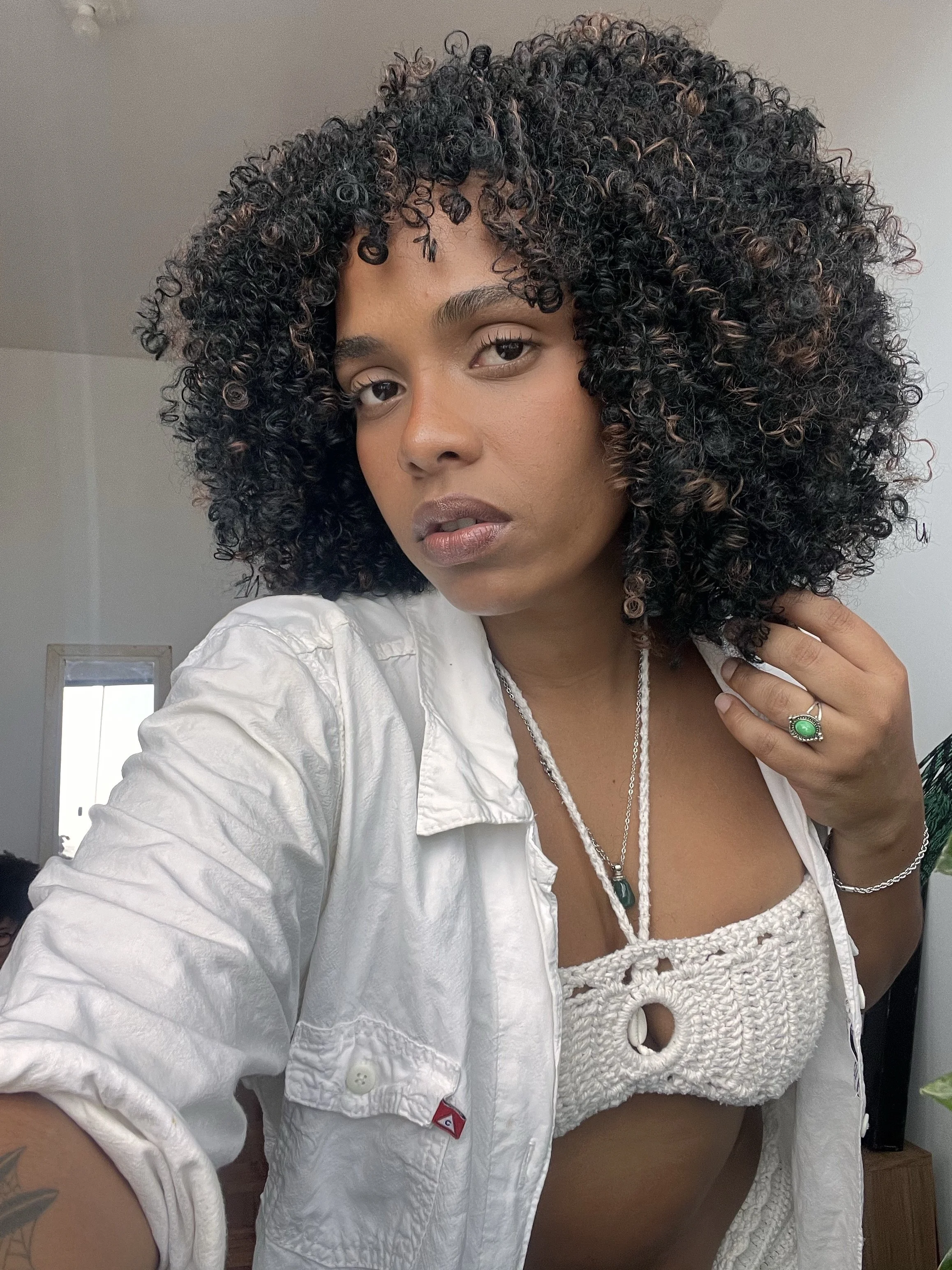 A woman with long, voluminous natural curls and a middle part, wearing a white button-down over a crochet top, showcasing how elongated curls add weight below the chin to balance a heart-shaped face.