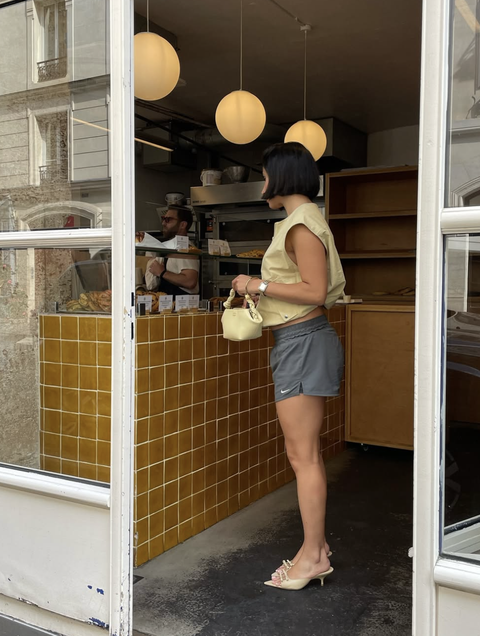A woman wearing casual grey Nike track shorts styled with an asymmetric pastel yellow knit top, a small matching cream handbag, and cream kitten heel mules.
