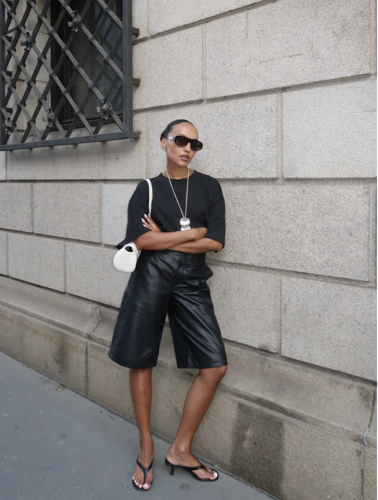 A woman wearing a black t-shirt and knee-length black leather Bermuda shorts paired with black kitten heel flip-flop sandals and a white shoulder bag.