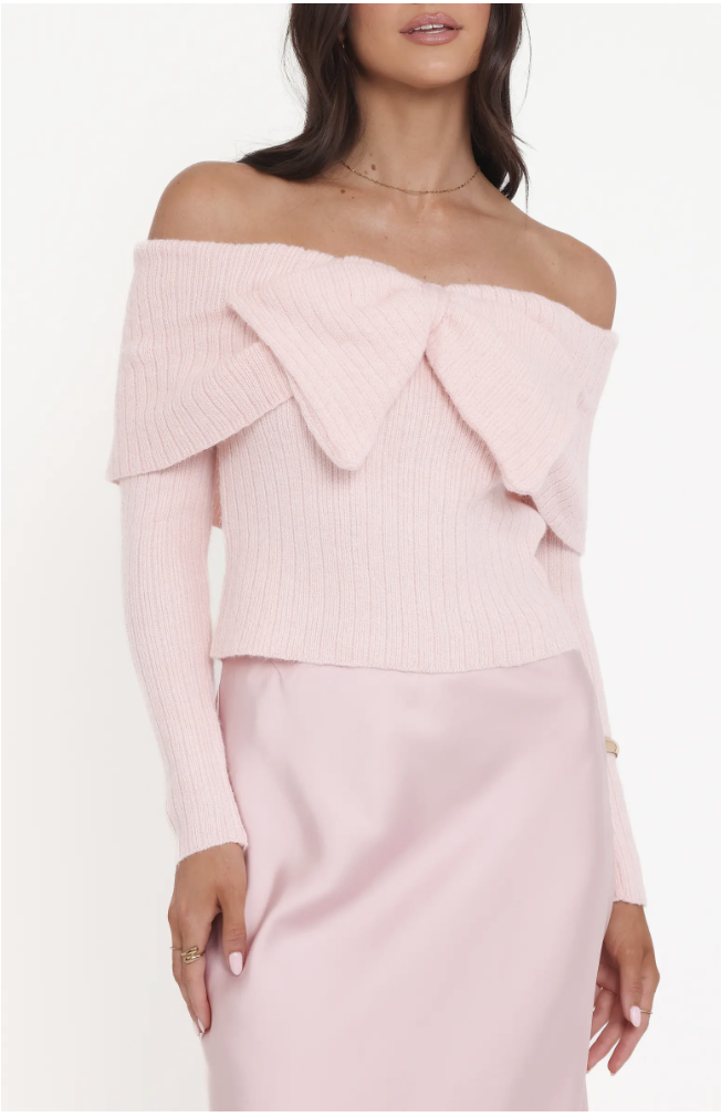 A woman wearing a light pink off-the-shoulder ribbed knit sweater with a layered fold-over neckline, paired with a matching pink satin skirt.