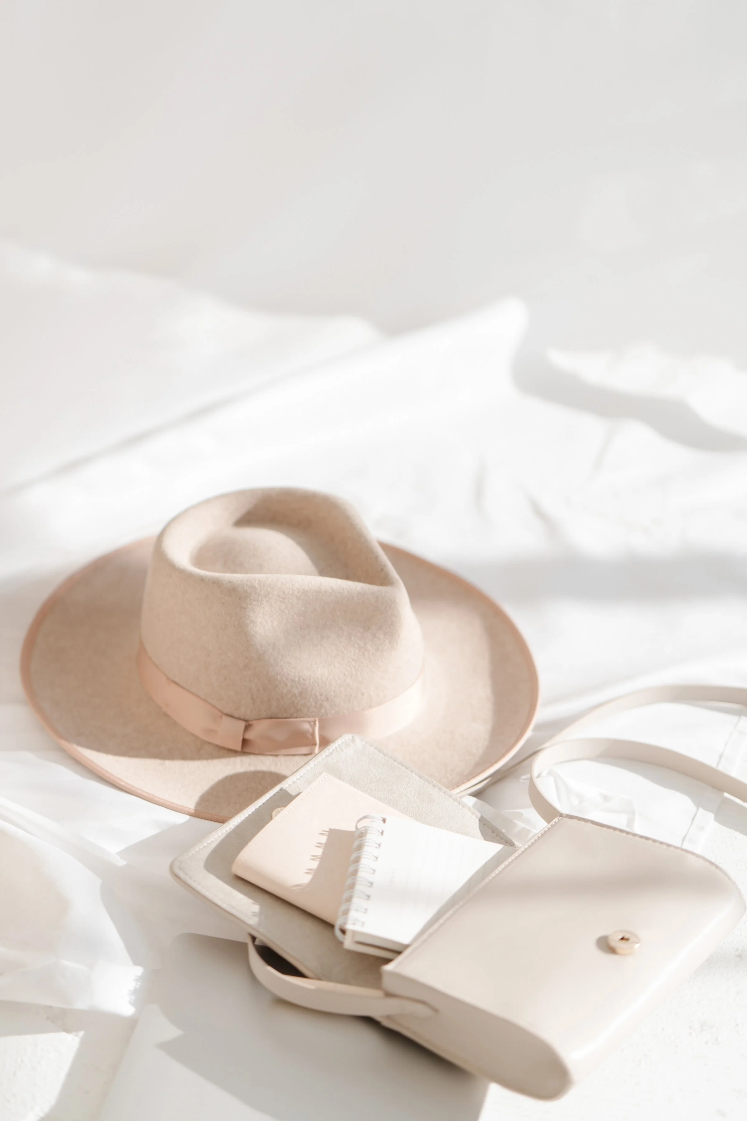 A beige wide-brimmed hat with a satin ribbon, a small white purse, and a notepad with a pen inside an open case, all resting on a white surface with soft natural lighting.