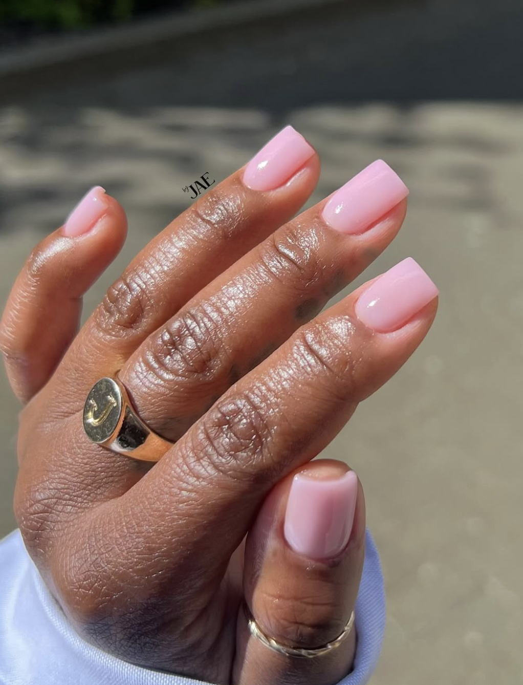 A close-up of a hand with short, square nails painted in a semi-sheer, milky pink polish with gold ring accents.