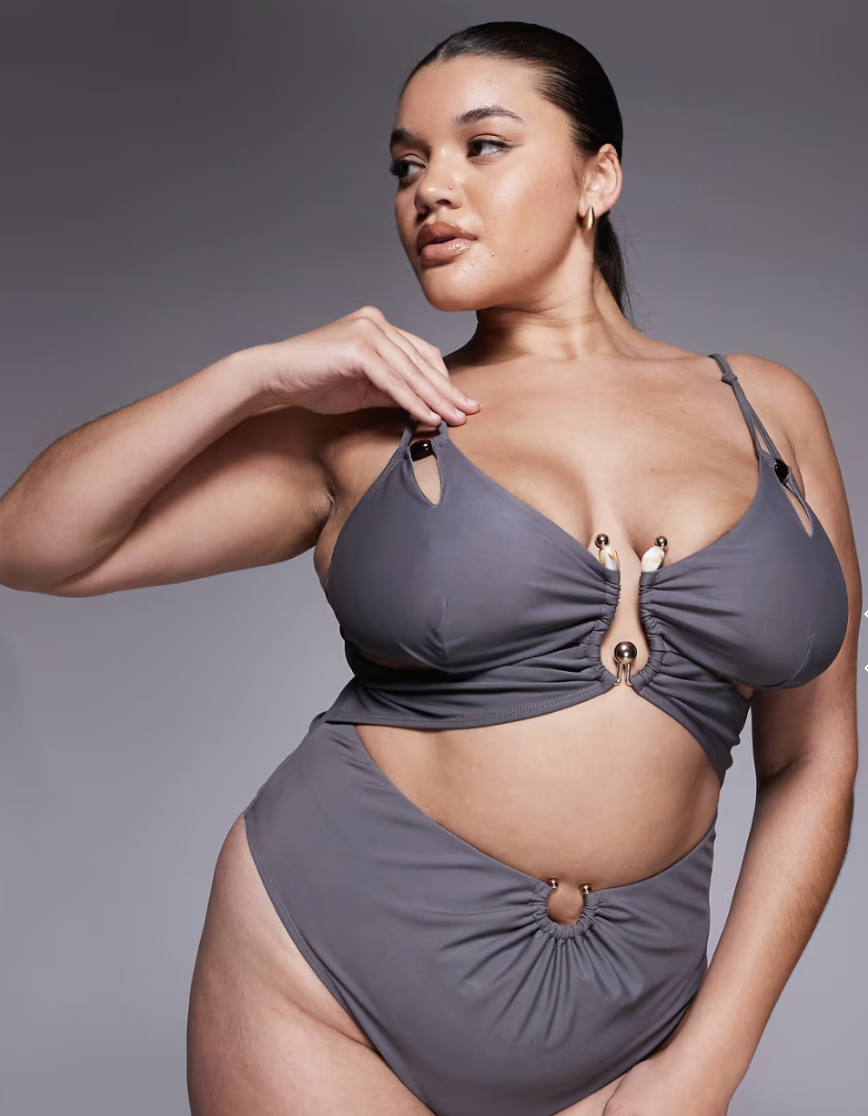 A woman wearing a grey one-piece swimsuit featuring an asymmetric cutout at the waist and a decorative silver ring detail.