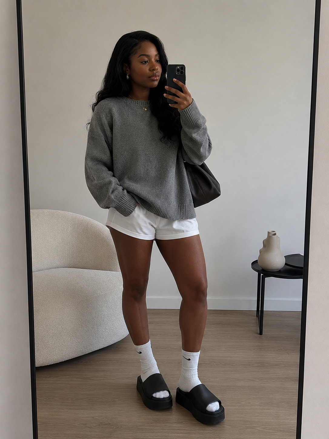 A woman taking a mirror selfie wearing an oversized grey crewneck sweater, white shorts, white athletic crew socks, and black platform slides, holding a black shoulder bag.