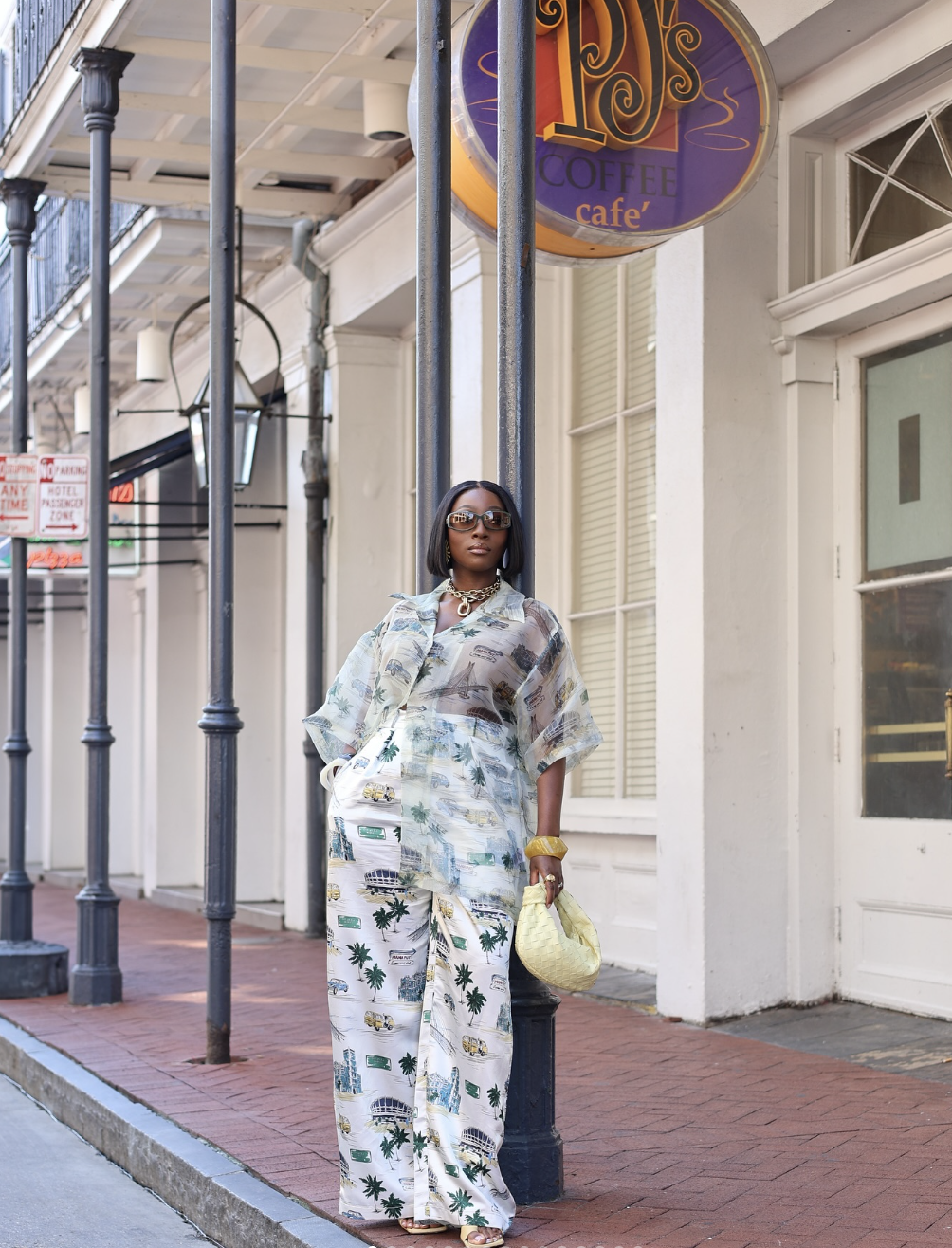 A woman standing on a city sidewalk under a café sign wearing a lightweight printed button-down shirt, oversized brown sunglasses, and a chunky gold chain necklace.
