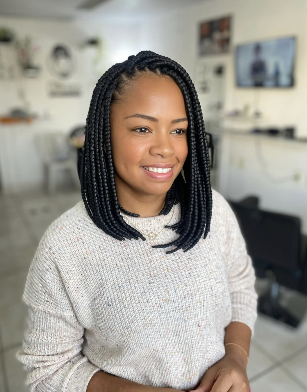 Close-up portrait of a smiling woman showcasing a neat, shoulder-length protective braided bob hairstyle, wearing a cozy cream knit sweater.