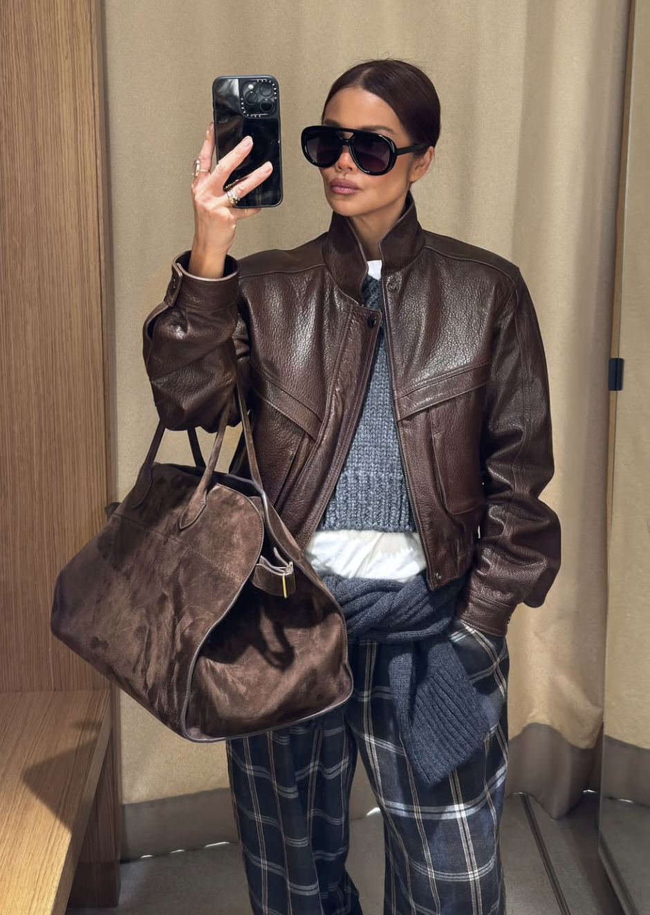 A woman wearing a chocolate brown leather bomber jacket layered over a grey knit sweater and blue plaid trousers, carrying a large brown suede tote bag.