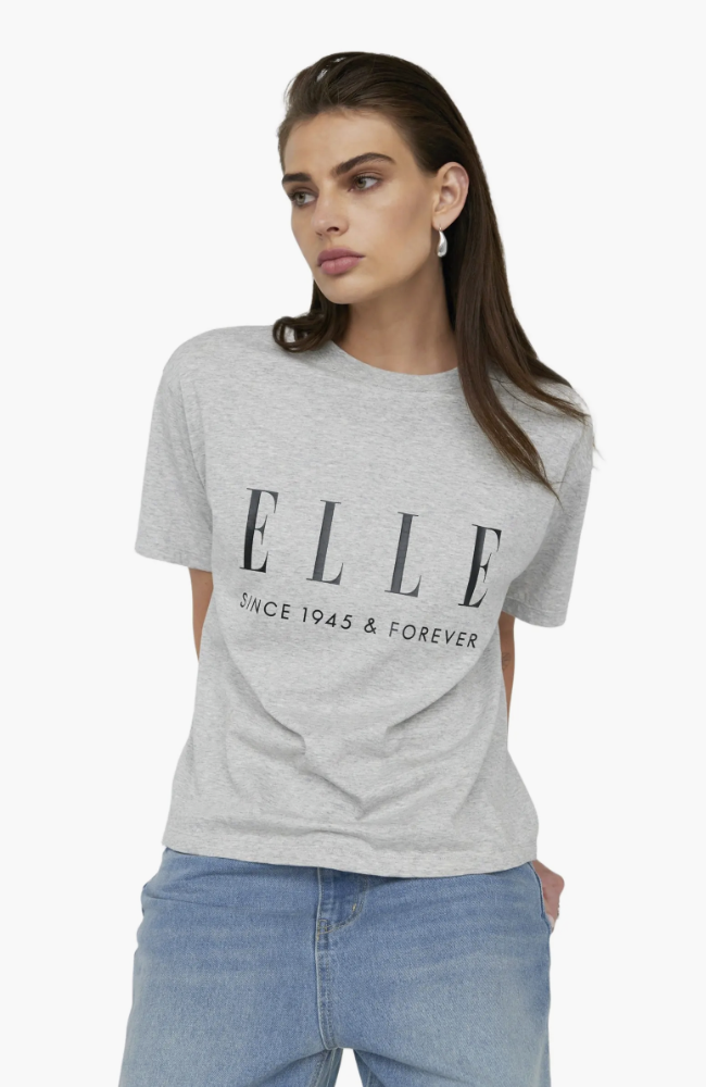 A woman wearing a grey "ELLE Since 1945 & Forever" graphic t-shirt with light wash blue jeans and silver earrings.