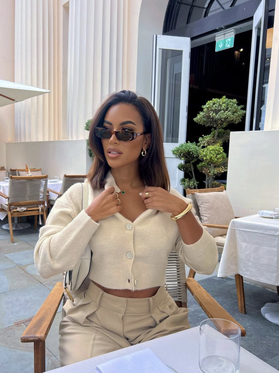 A woman wearing a quiet luxury neutral outfit featuring a cropped cream button-up cardigan paired with high-waisted beige trousers and gold jewelry.