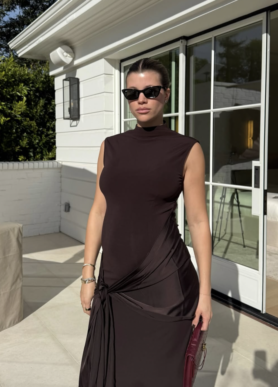 Sofia Richie wearing a chocolate brown sleeveless maxi dress with a draped knot detail and black sunglasses, epitomizing the quiet money aesthetic.