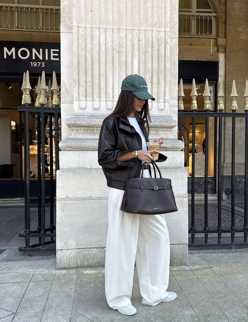 A woman wearing a polished city outfit featuring an oversized brown leather jacket, white wide-leg trousers, a deep espresso tote bag, and a green baseball cap.