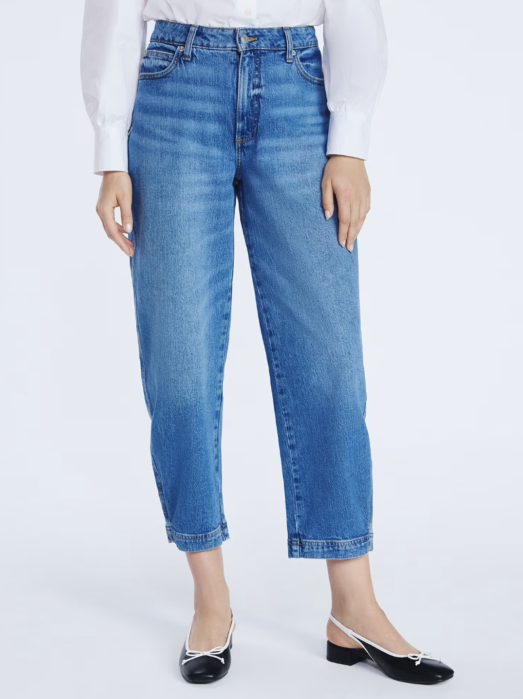 A person wearing mid-rise wide hem barrel jeans in a classic blue wash, paired with white bow-detail ballet flats, highlighting the architectural tapered leg and cropped ankle length.
