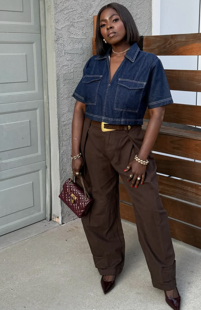 A person wearing a dark wash denim short-sleeve button-down shirt tucked into chocolate brown wide-leg trousers with a tan leather belt and a quilted burgundy handbag.