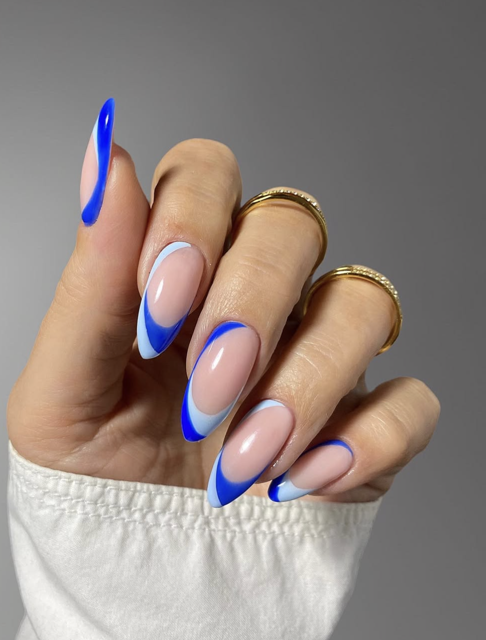 Long, oval-shaped nails with a translucent base coat featuring an abstract, multi-layered French tip design in vibrant cobalt and soft pastel blue waves.