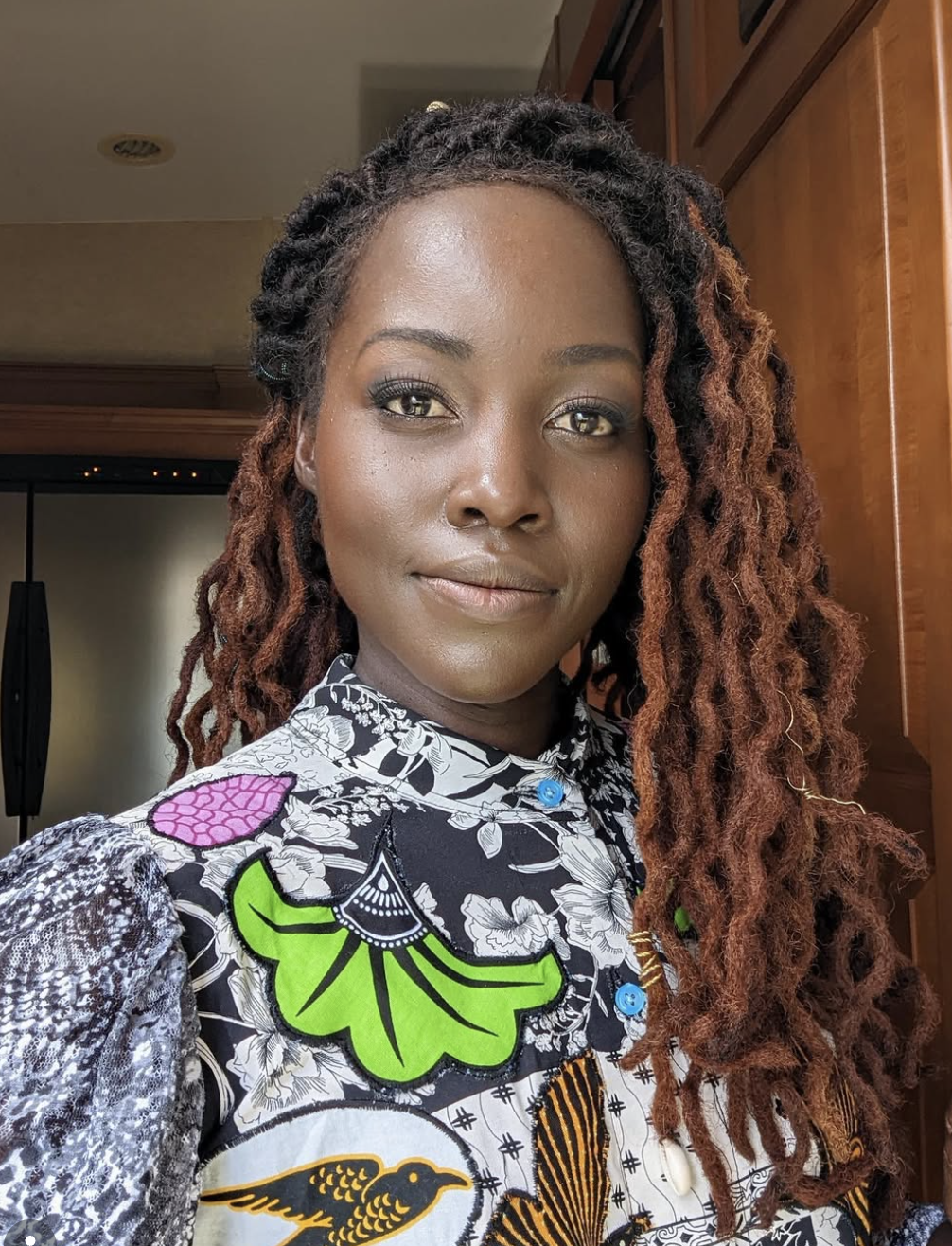 Lupita Nyong'o wearing long, mahogany-brown textured locs and a vibrant floral print blouse with green and purple accents.