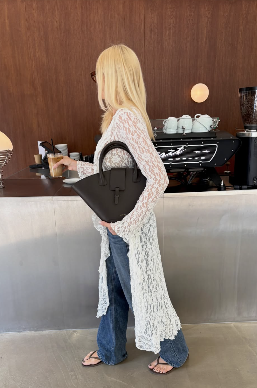 A woman wearing a floor-length white lace duster cardigan over wide-leg blue jeans and casual black flip-flop sandals.