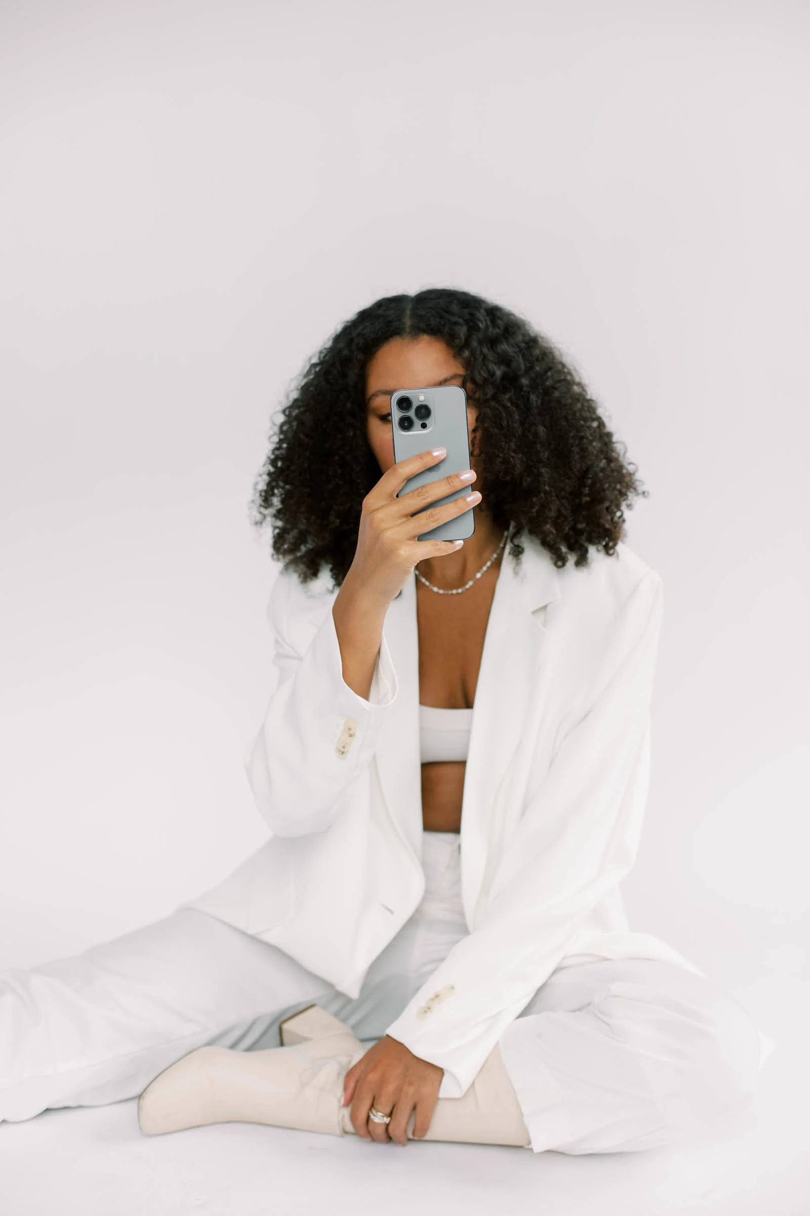 Woman with curly hair taking a selfie with her phone, dressed in a white suit and seated on the floor against a plain white background.