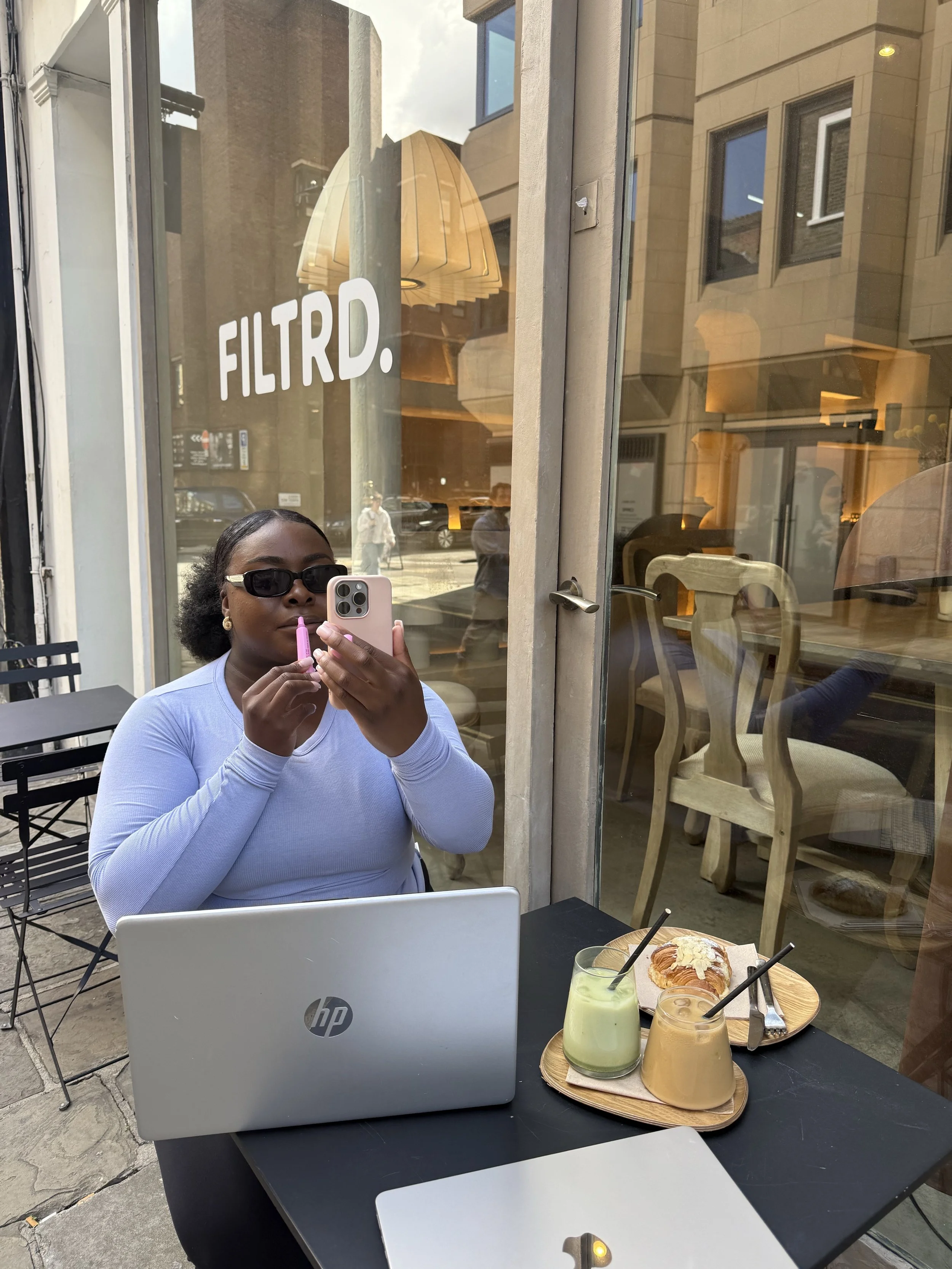 A woman sitting at an outdoor cafe table applying lip gloss while looking into her phone camera, with a laptop and matcha latte in the foreground.