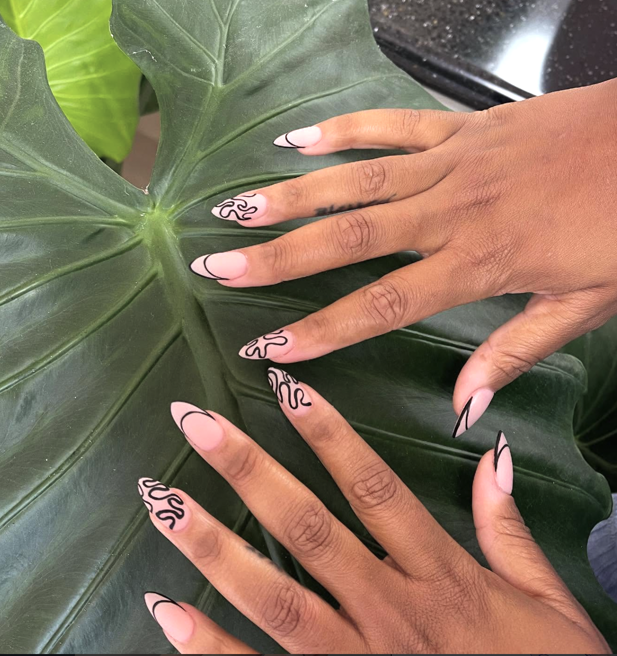 Long almond-shaped nails with a sheer nude base and hand-painted black swirl patterns that mimic a French tip shape, featuring white accents and a single solid white tip on the index finger.