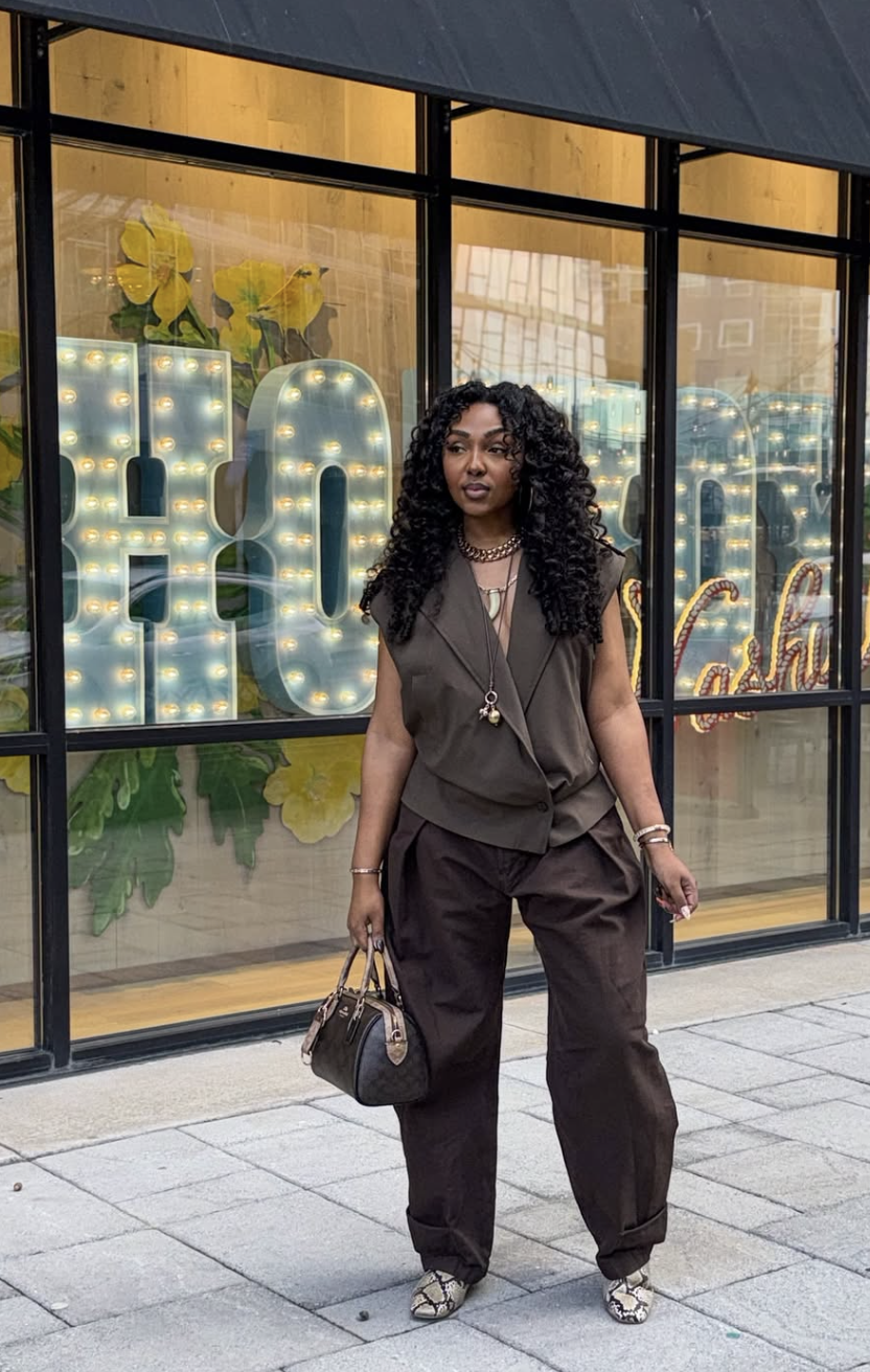 A woman with long, curly black hair wearing a dark brown sleeveless tailored vest and matching wide-leg pleated trousers, carrying a brown patterned handbag, and wearing snakeskin-print shoes.