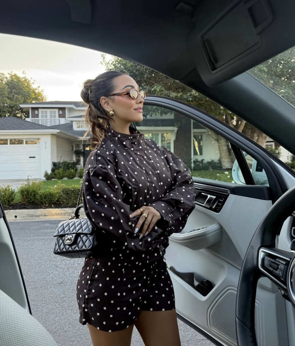 A woman leaning against a car door wearing a chocolate brown and white polka dot matching set, featuring a high-neck long-sleeve blouse and a micro-mini skirt, styled with a black quilted handbag and oval sunglasses.