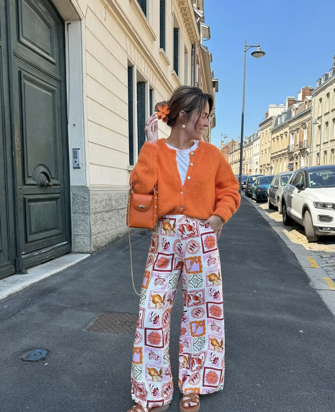 A woman wearing a monochromatic orange outfit featuring a bright tangerine cardigan layered over a white top and high-waisted orange patterned wide-leg trousers.