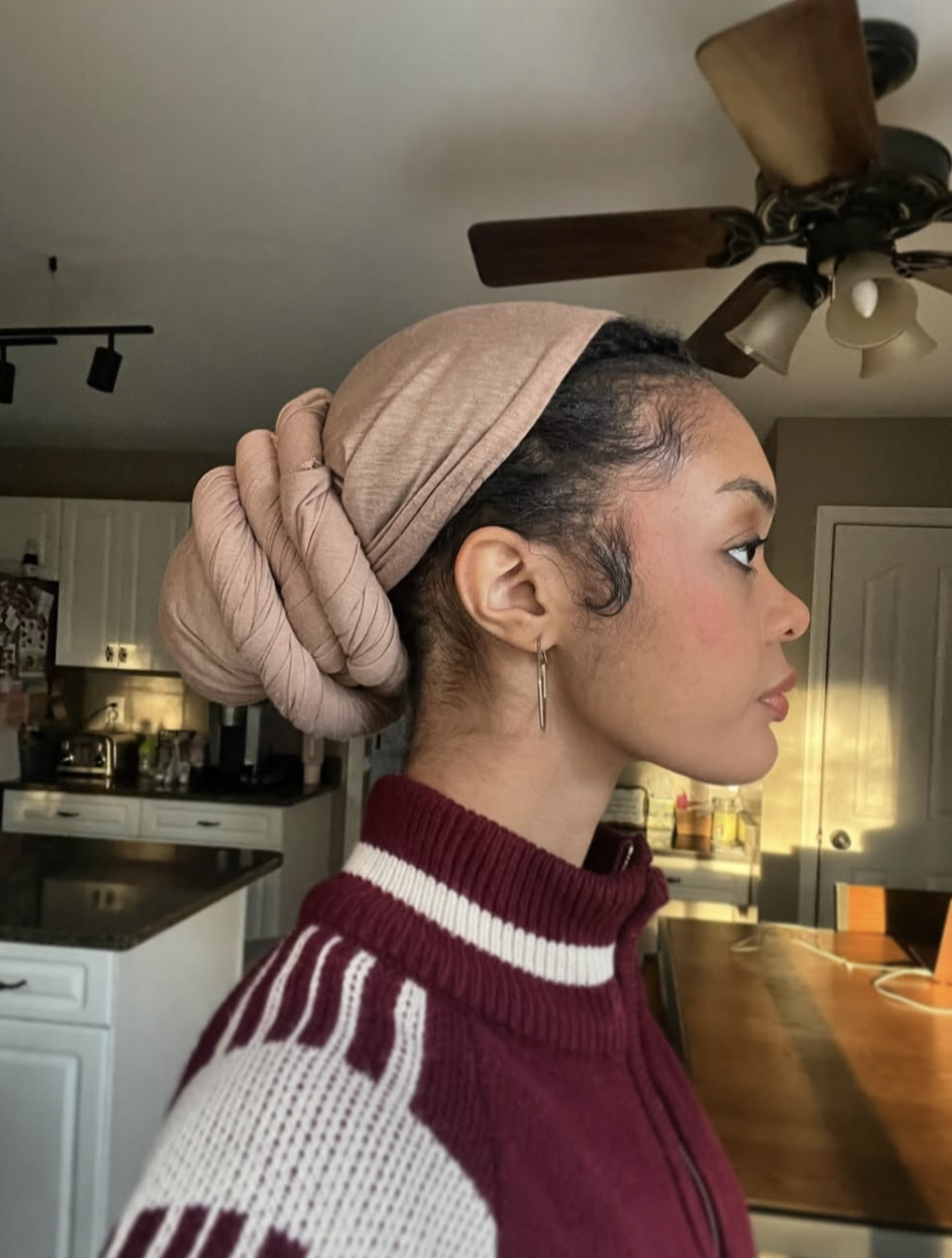 A close-up profile shot of a woman wearing a mauve-pink silk scarf wrapped into a full protective head wrap style with a neat top knot, paired with a burgundy and white ribbed knit zip-up sweater and silver hoop earrings.