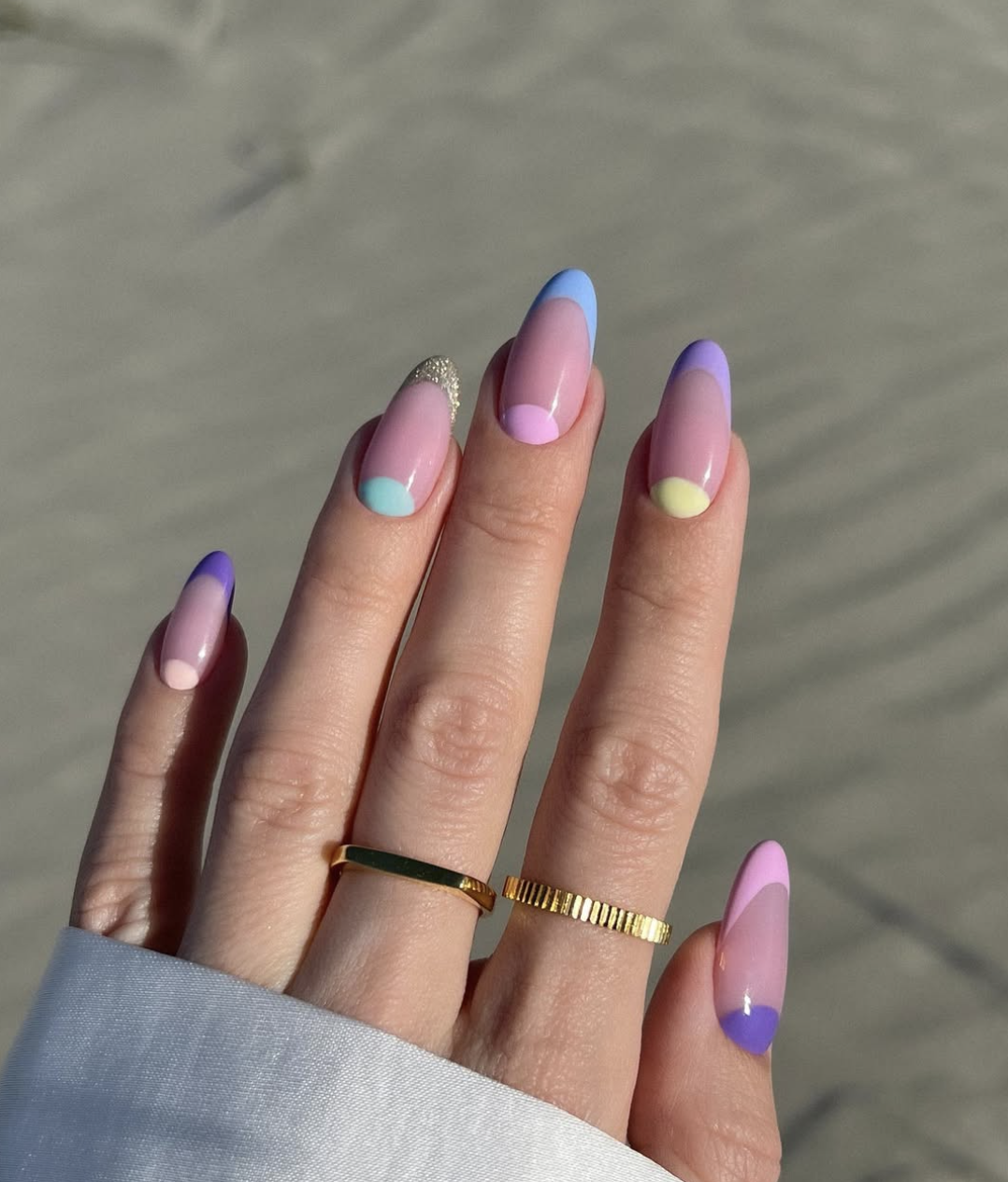Close-up of a hand showcasing a creative summer manicure featuring a mix of lavender and periwinkle French tips combined with colorful negative-space half-moon designs at the cuticles, styled with simple gold bands.