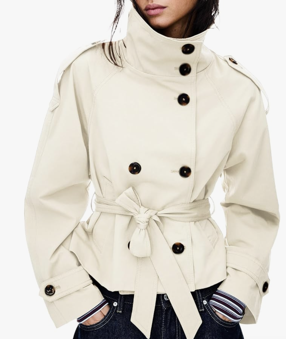 A woman wearing a cropped cream-colored trench coat with dark buttons, styled with a high-waisted belt and a sweater draped over her shoulders.