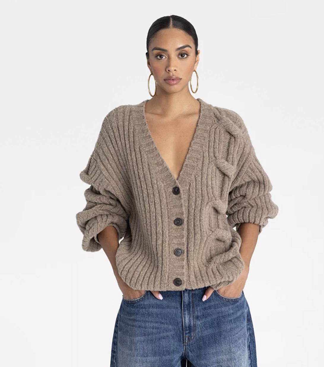 A woman wearing a taupe cable-knit cardigan with oversized buttons tucked into light blue jeans.