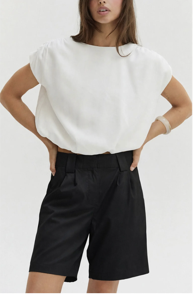 A close-up view of a woman styling high-waisted, structured black tailored shorts with sharp pleats, paired with an everyday white cap-sleeve top.