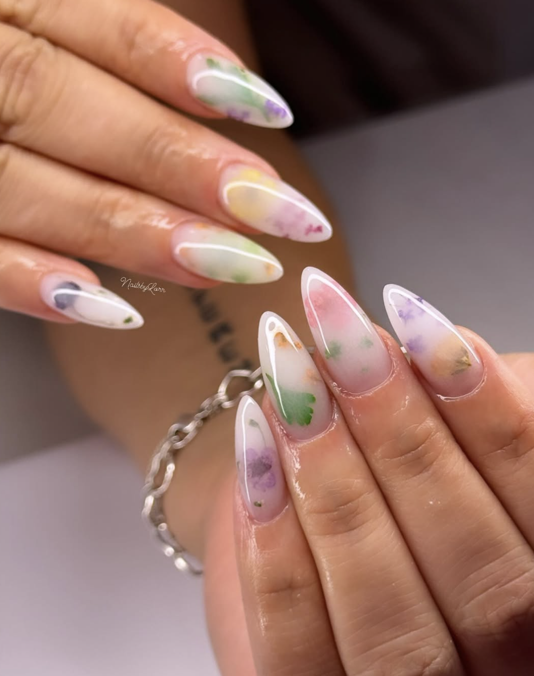 Long almond-shaped nails with a translucent white milk bath finish featuring embedded purple and green dried botanical accents.