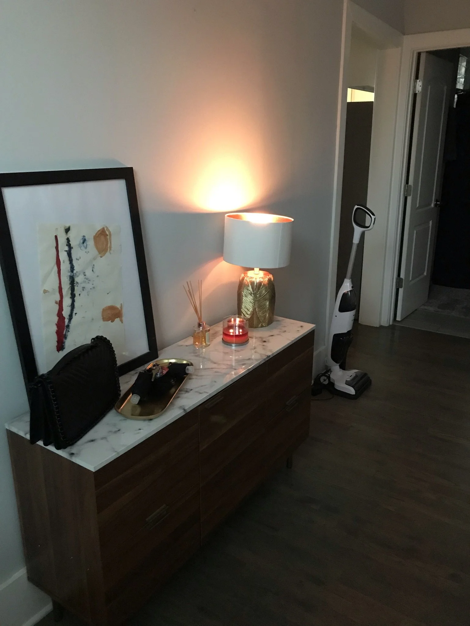 Apartment entryway with a walnut sideboard, marble top, glowing table lamp, abstract art and a cordless vacuum by the door