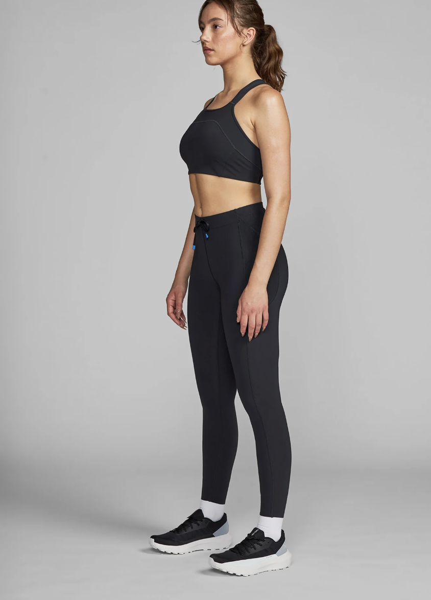 A side-view detail of the Athleta Rainier High Rise Legging in black, showcasing the functional zip pocket on the thigh and the supportive, high-waisted compression fit styled with white running sneakers.