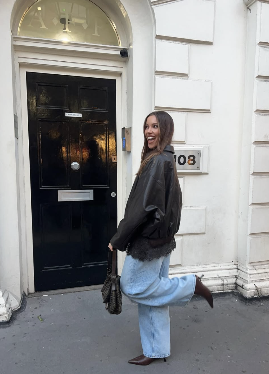 A woman posing in front of a black door wearing baggy light blue jeans, a black oversized leather bomber jacket with lace trim, and chocolate brown pointed-toe heels.