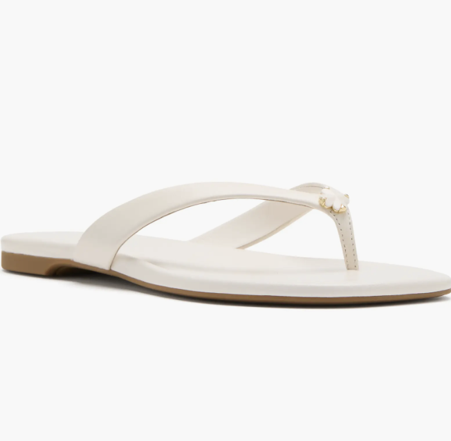 A close-up side profile of a minimalist white leather flat flip flop sandal with a circular metal detail on the strap.
