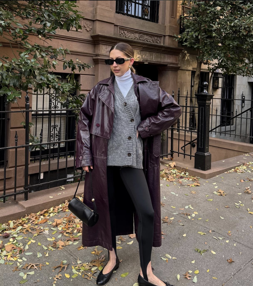 Woman wearing black stirrup leggings with a long plum-colored leather trench coat, a grey button-up cardigan, and black leather ballet flats.