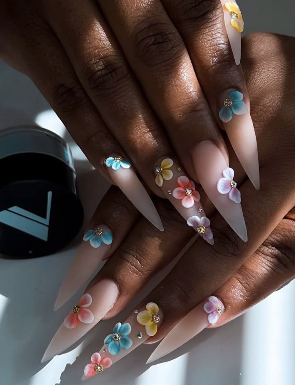 Close-up of a hand showcasing a detailed vacation manicure featuring 3D sculpted flowers with yellow and blue petals and tiny gold bead centers on long, natural-shaped nails.