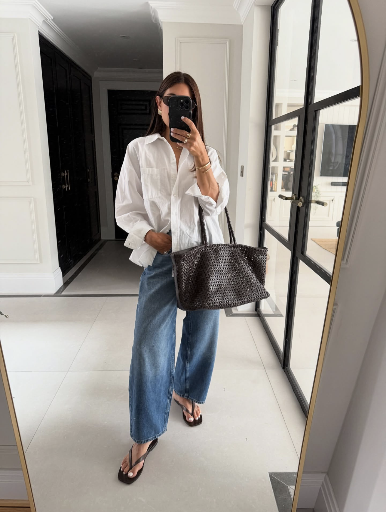 A woman wearing an oversized white button-down shirt tucked into medium-wash barrel-leg jeans, accessorized with a large black woven tote bag and black flip-flop sandals.