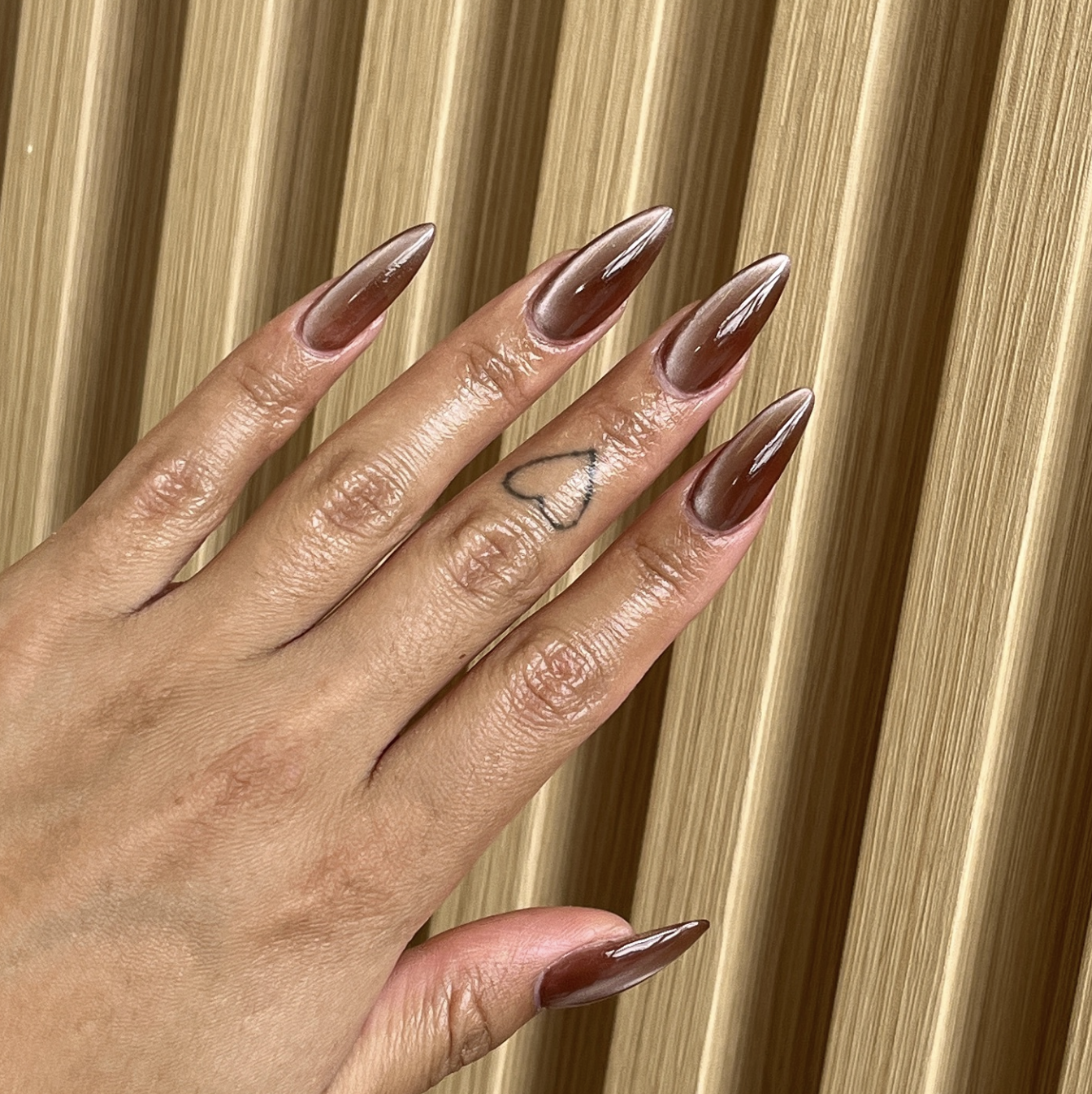 A close-up of long almond-shaped nails featuring a reflective metallic brown chrome finish with a small finger tattoo.