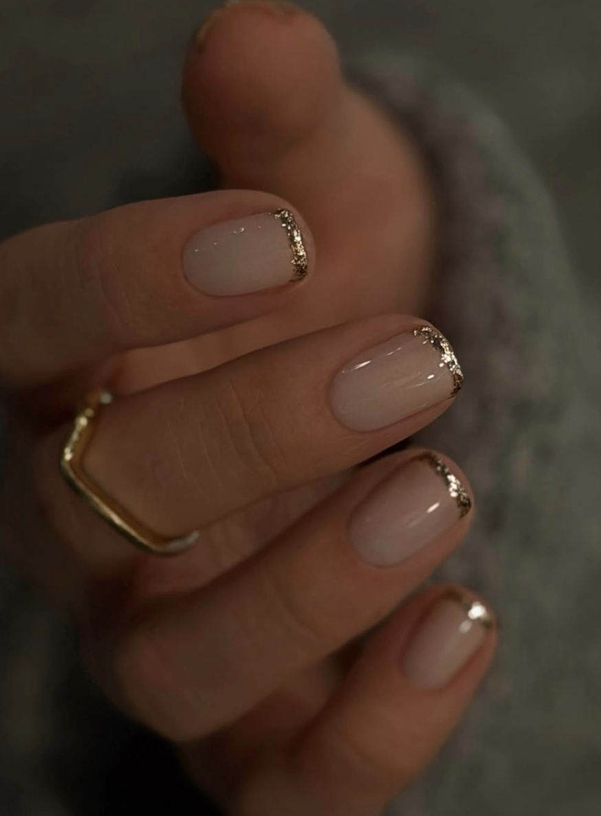 A close-up of short nails with a sheer nude base accented with metallic gold foil flecks along the cuticles.