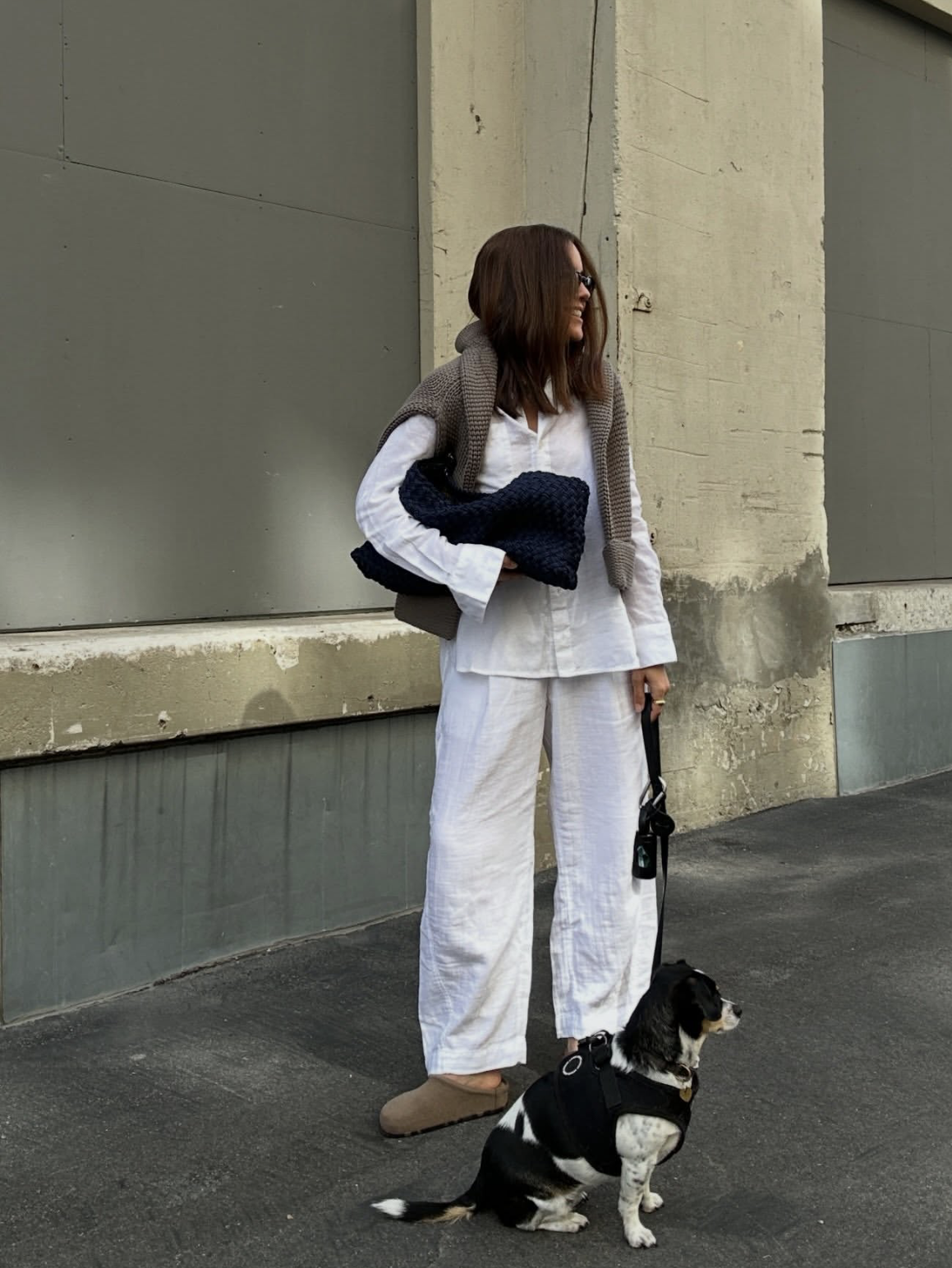 A chic street style look featuring a woman in a matching white linen button-down shirt and wide-leg linen pants, styled with tan suede clogs and a brown knit sweater draped over her shoulders.