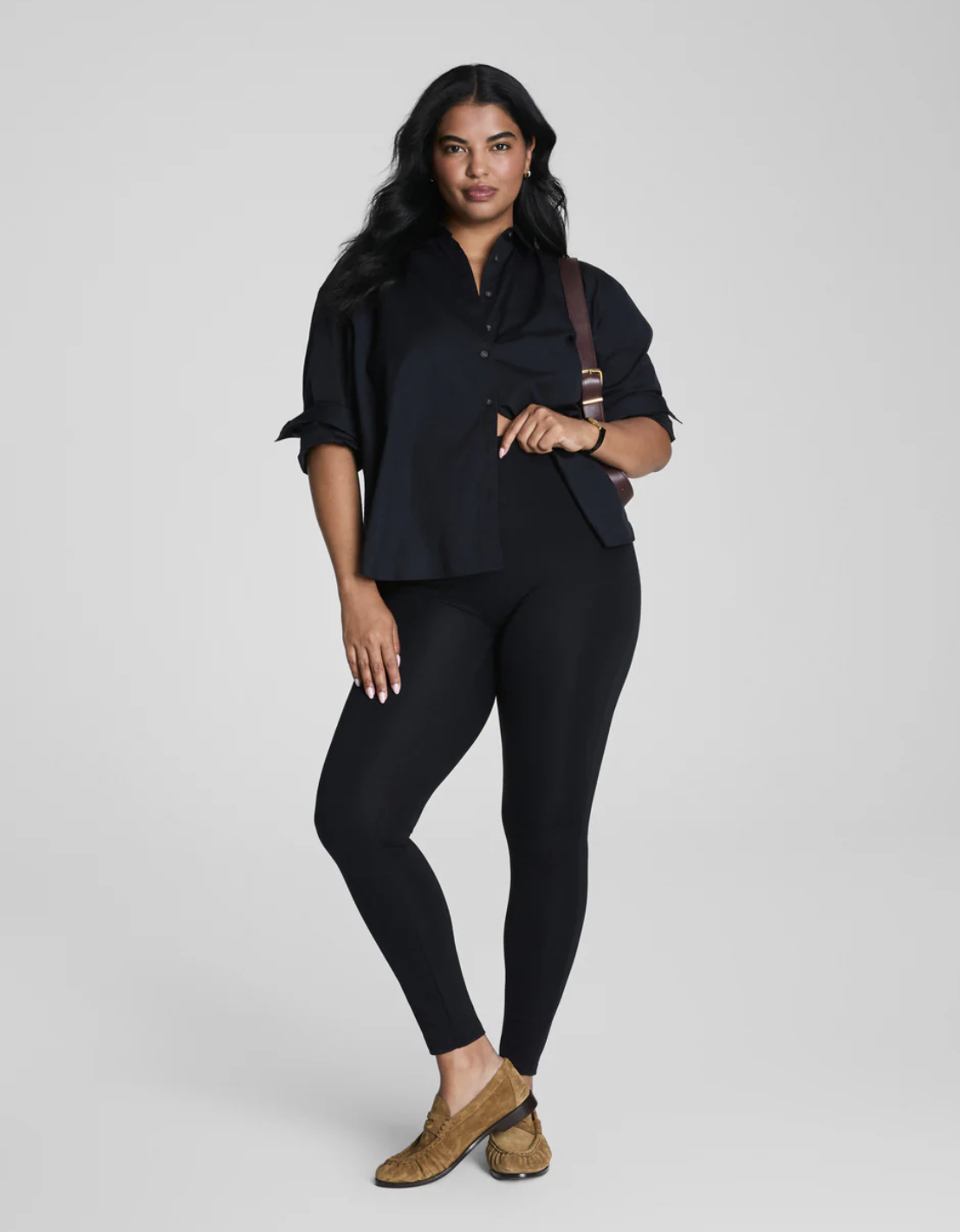 A plus-size model wearing black high-waisted Universal Standard Roya leggings with a black button-down shirt and brown ankle boots, demonstrating a polished way to style performance leggings for everyday life in 2026.