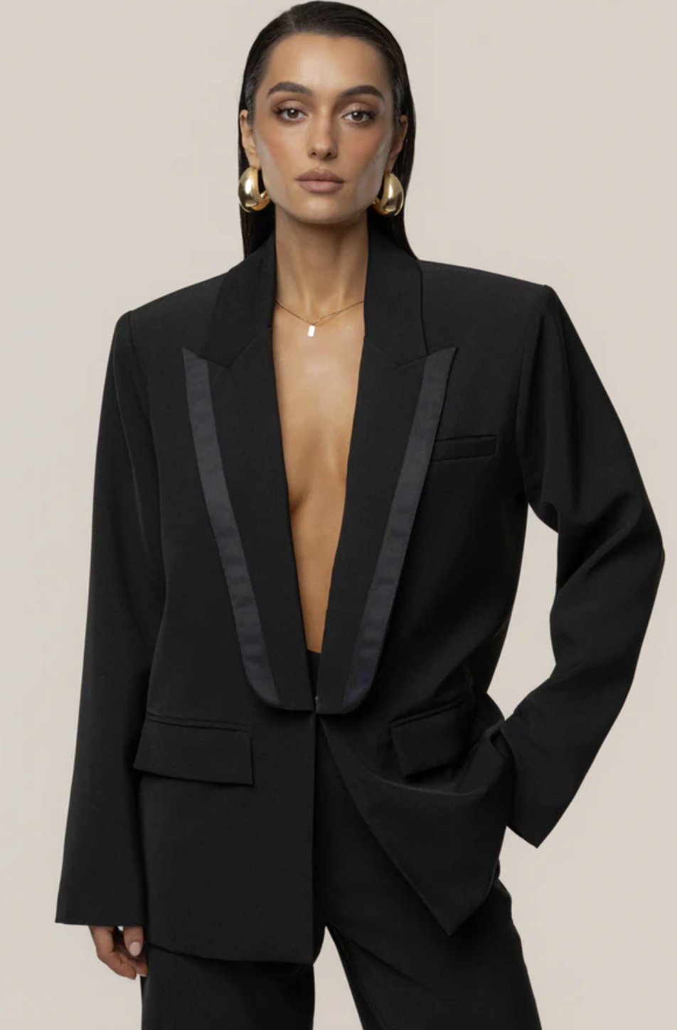 A woman wearing a black structured tuxedo-style blazer with oversized gold earrings and a slicked-back hairstyle to demonstrate a polished way to style baggy jeans.