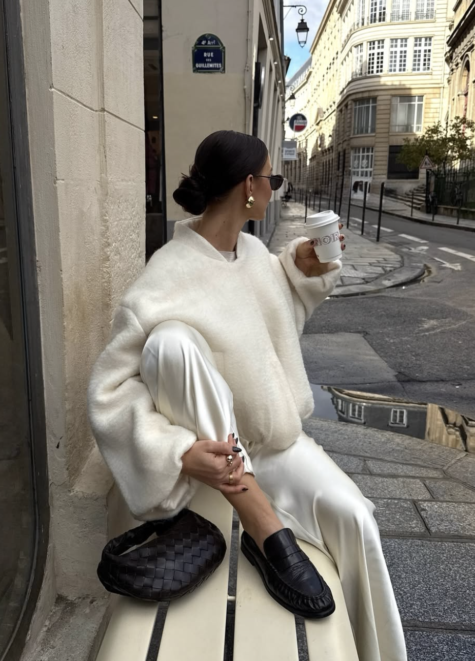 A woman wearing a cozy monochromatic white and cream outfit featuring an oversized ribbed sweater, white trousers, and gold hoop earrings, holding a coffee cup while walking in a city.