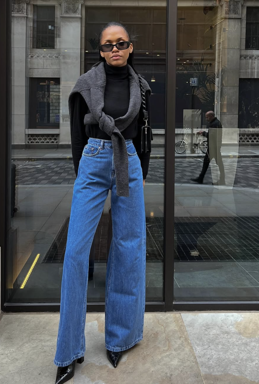 A woman wearing high-waisted wide-leg jeans, a black turtleneck, pointed-toe heels, and a gray sweater draped over her shoulders.