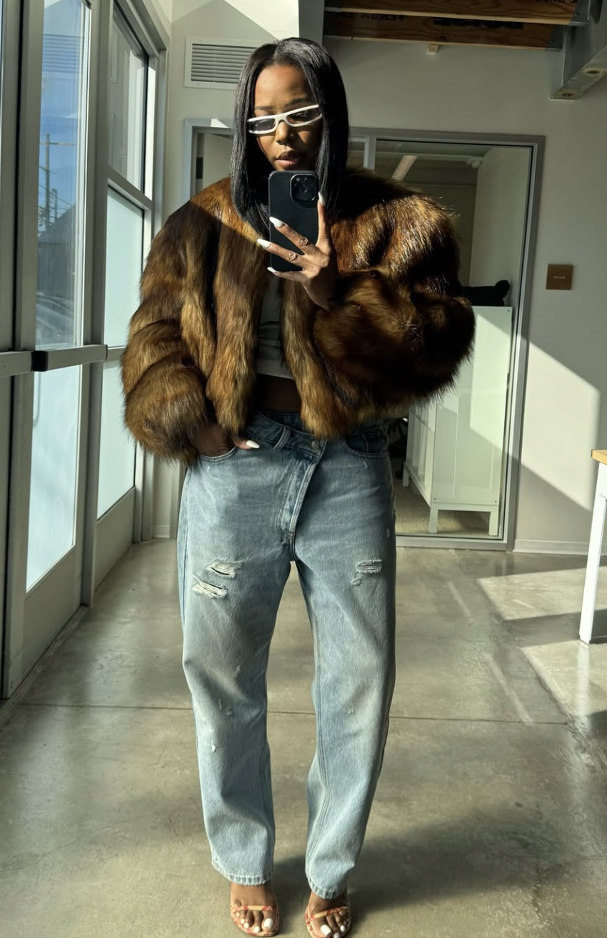 A woman wearing light blue distressed jeans with a criss-cross waistband, a white crop top, and a voluminous brown faux fur jacket.