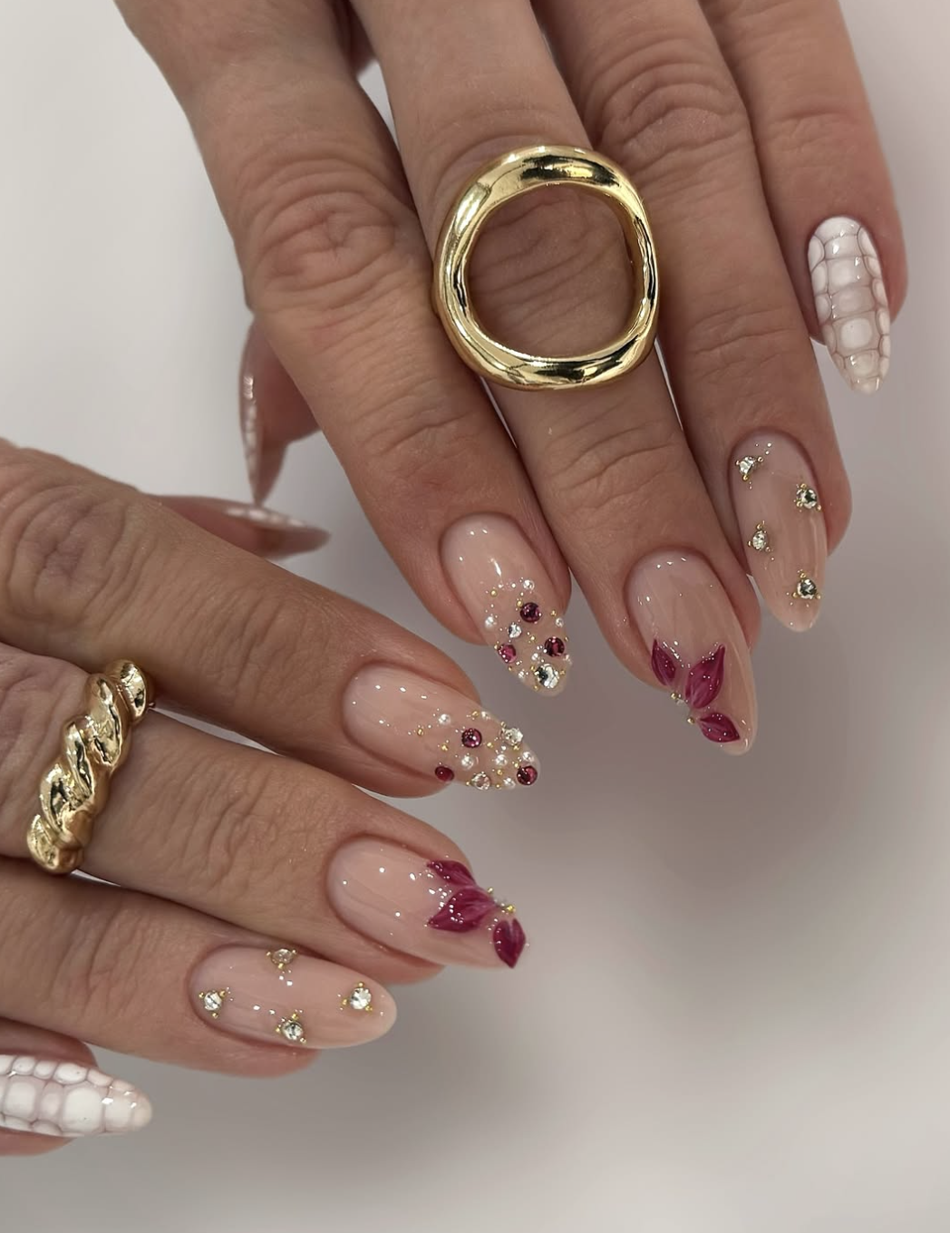 A high-feminine luxury manicure by @__mknails featuring almond-shaped nails with 3D white floral textures, tiny gold studs, and deep burgundy petal details on a sheer nude base.